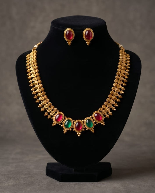 Gold necklace with red and green gemstones and matching earrings on a black stand against a gray background